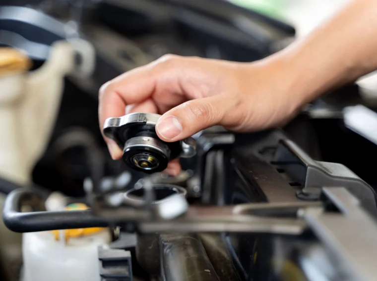 A person’s hand is removing a radiator cap from a car engine compartment, preparing for cooling system repair.