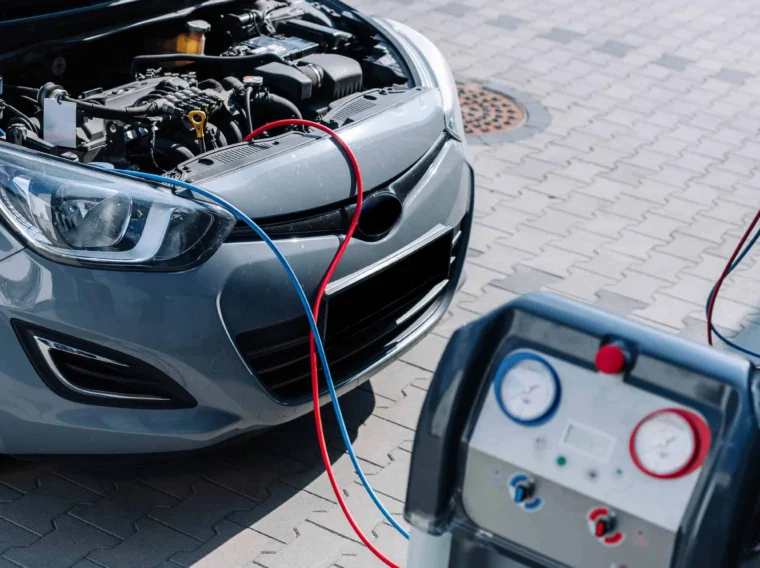A car with its hood open is connected by hoses to an air conditioning service machine for maintenance or air conditioning recharge on a paved surface.