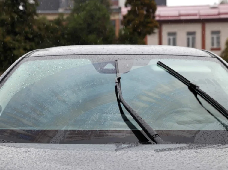 A car windshield with rain droplets is being cleared by two windshield wiper blades in motion. Buildings and trees are visible in the background.