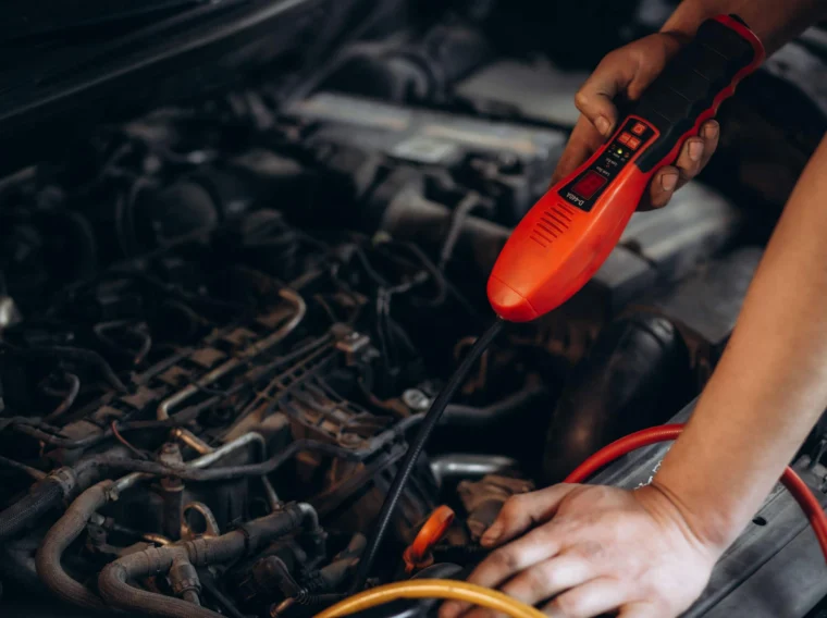 A person uses a diagnostic tool under the hood of a car, checking engine components with one hand while holding the device with the other during an AC Recharge.