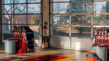 An empty auto repair garage with equipment, tool racks, and a shop vacuum—perfect for Battery Services or Car Battery replacement—lit by sunlight through large glass doors.