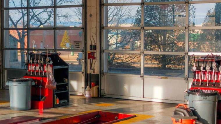An empty auto repair garage with equipment, tool racks, and a shop vacuum—perfect for Battery Services or Car Battery replacement—lit by sunlight through large glass doors.