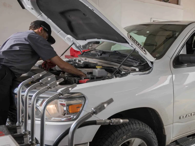 A mechanic inspects under the hood of a white Ram truck, possibly checking Battery Services, while a person sits inside the vehicle at an automotive repair shop.