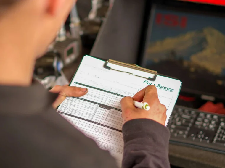 A person holding a pen fills out a form on a clipboard, standing next to a computer and automotive diagnostic equipment for battery services.