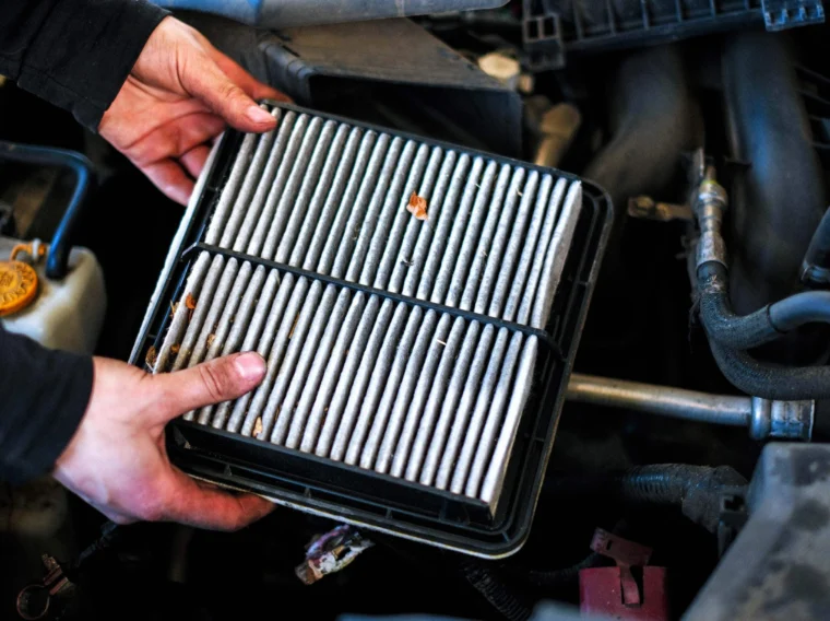 A person holds a rectangular car air filter above an engine bay, showing some dirt and debris trapped in the filter—a clear sign it's time to consider professional Filter Services.
