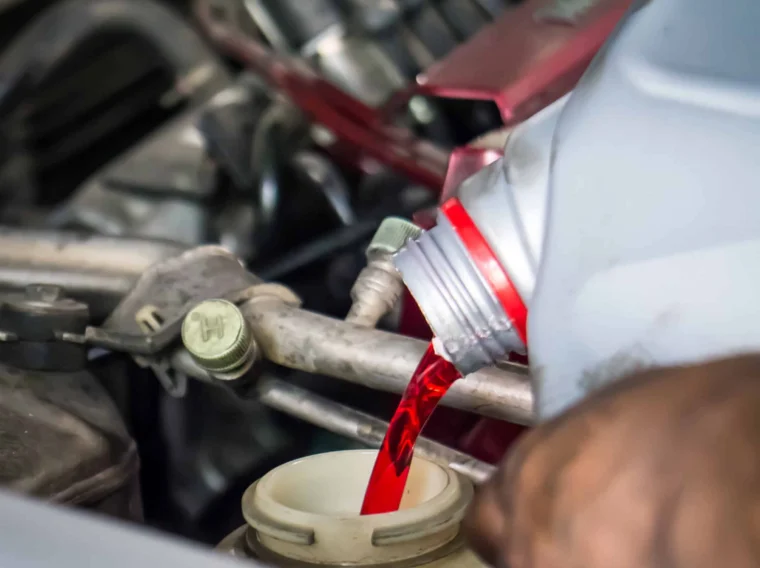 A person pours red engine oil or transmission fluid from a plastic container into a car’s engine compartment.