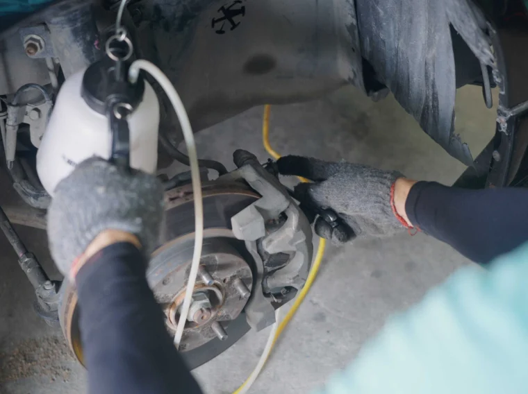 Person wearing gloves uses a brake bleeder tool on a car brake caliper with the wheel removed in a garage setting.