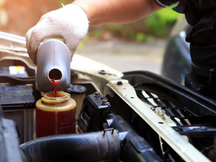 A person wearing a white glove pours red transmission fluid from a plastic bottle into a car’s power steering system under the hood.