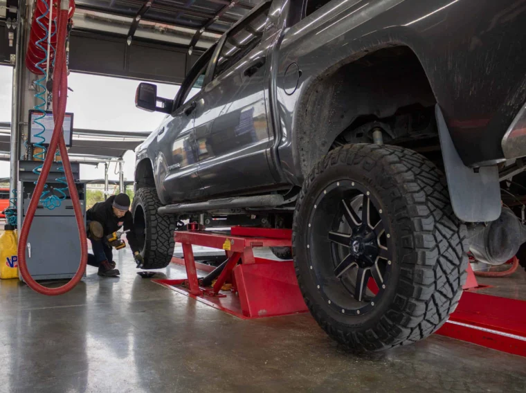 A mechanic inspects a black pickup truck lifted on a red hydraulic platform inside a garage, preparing for tire installation and other tire services.