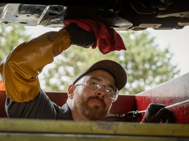 A mechanic wearing safety glasses and gloves works under a vehicle, holding a red cloth and inspecting the underside while performing a drivetrain fluid replacement.