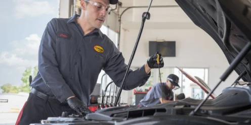 A mechanic in uniform checks the oil dipstick of a car in a service garage, with another worker visible in the background.