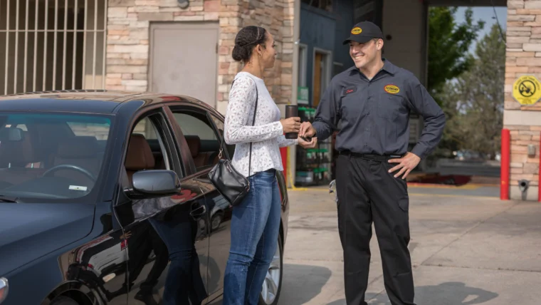 A woman stands next to a car, receiving keys from a uniformed mechanic outside an auto repair shop.